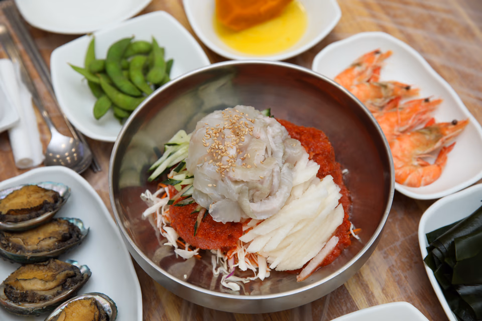 Chopsticks lifting noodles and raw fish from a bowl of mulhoe