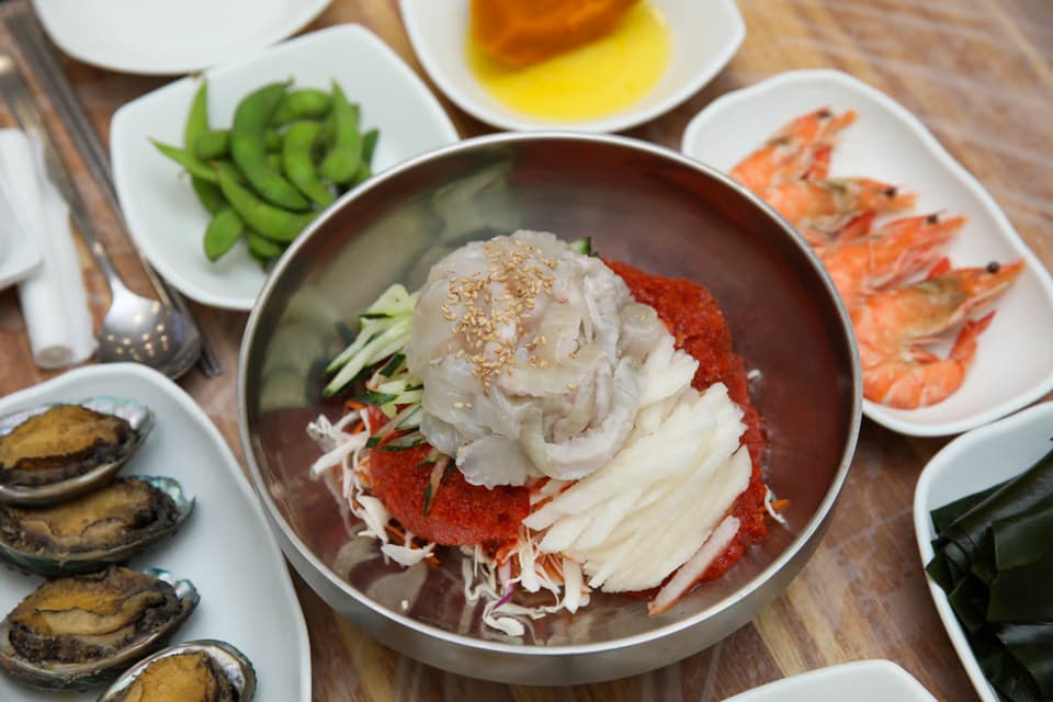 Chopsticks lifting noodles and raw fish from a bowl of mulhoe