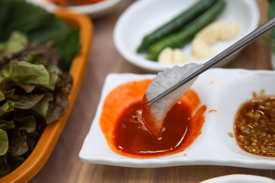 젓가락으로 두툼한 흰살 회 한 점을 집어 올린 장면, 뒤로 고급 모둠회 접시가 보이는 상