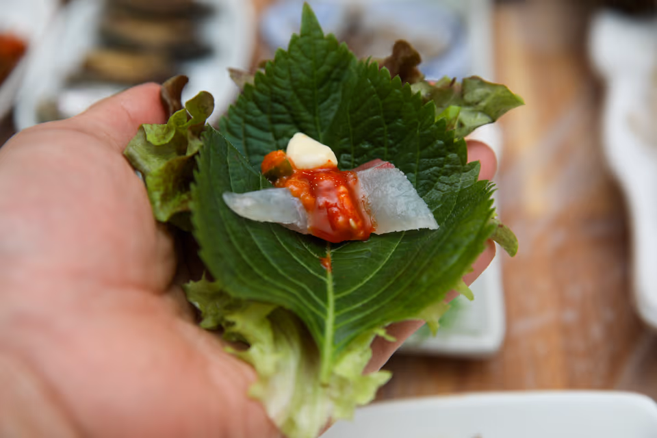 손에 든 깻잎과 상추 위에 흰살 회 한 점과 초장, 마늘을 올린 회 쌈