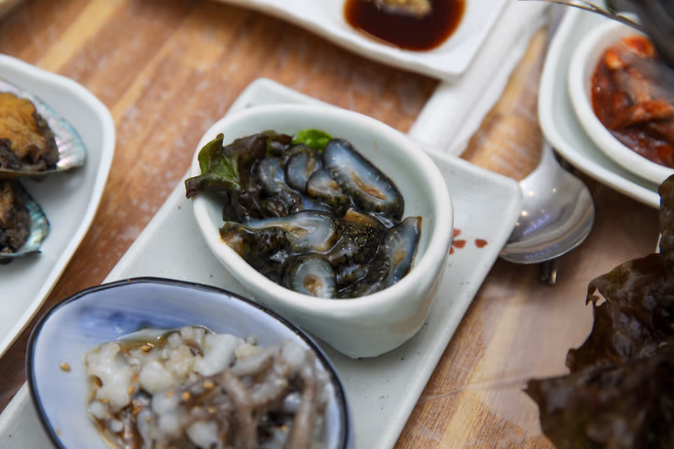 Dark sea cucumber sashimi in a white dish, with a sesame-sprinkled octopus bowl in front and abalone in the shell to the left