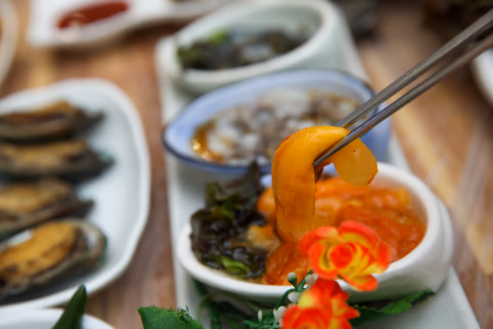 젓가락으로 주황빛 멍게 한 점을 집어 올린 장면, 뒤로 낙지 그릇과 해삼 종지, 왼쪽에 전복 접시