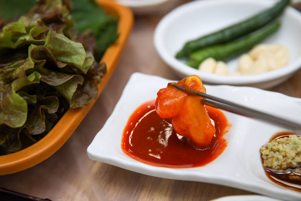 A piece of raw shellfish lifted on chopsticks after a generous dip in the chili paste dish, with a lettuce basket and chili pepper and garlic dishes behind