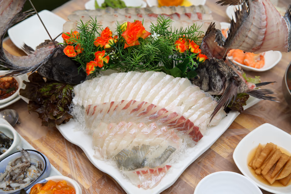 큰 흰 접시에 흰살 회를 둥글게 담고 우럭과 도미 장식, 꽃 장식을 올린 고급 모둠회, 옆에 반찬 종지들