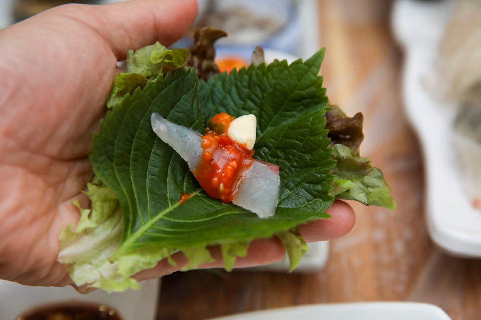 手上拿著紫蘇葉包,裡面是白肉生魚片、醋辣醬和一瓣大蒜,後面桌上擺著生魚片的盤子