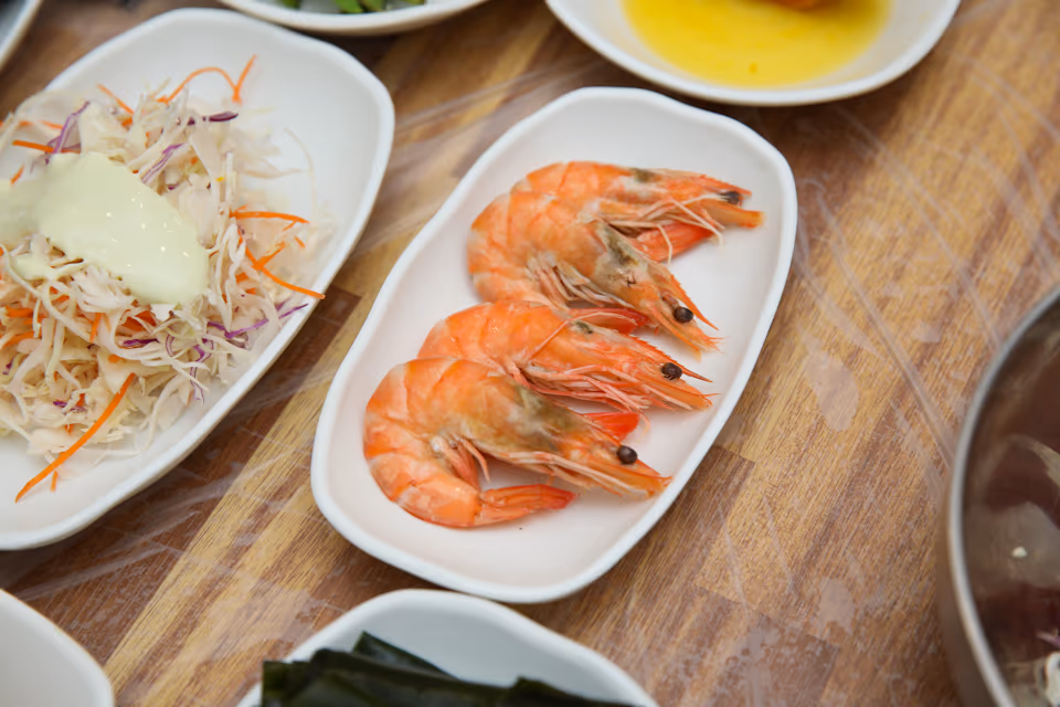 껍질째 익힌 새우 네 마리가 흰 접시에 가지런히 놓인 실사진, 옆에 채소 샐러드 종지가 보이는 상차림
