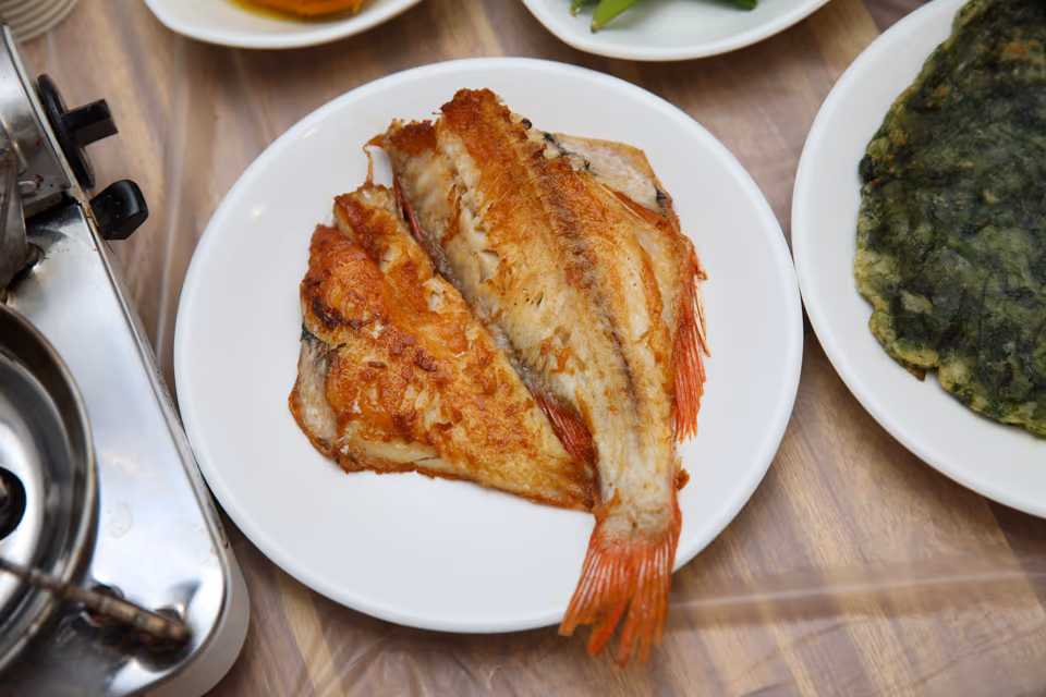 노릇하게 구운 생선이 흰 접시에 겹쳐 담긴 모듬생선구이 실사진, 옆으로 부추전 접시가 보이는 상차림